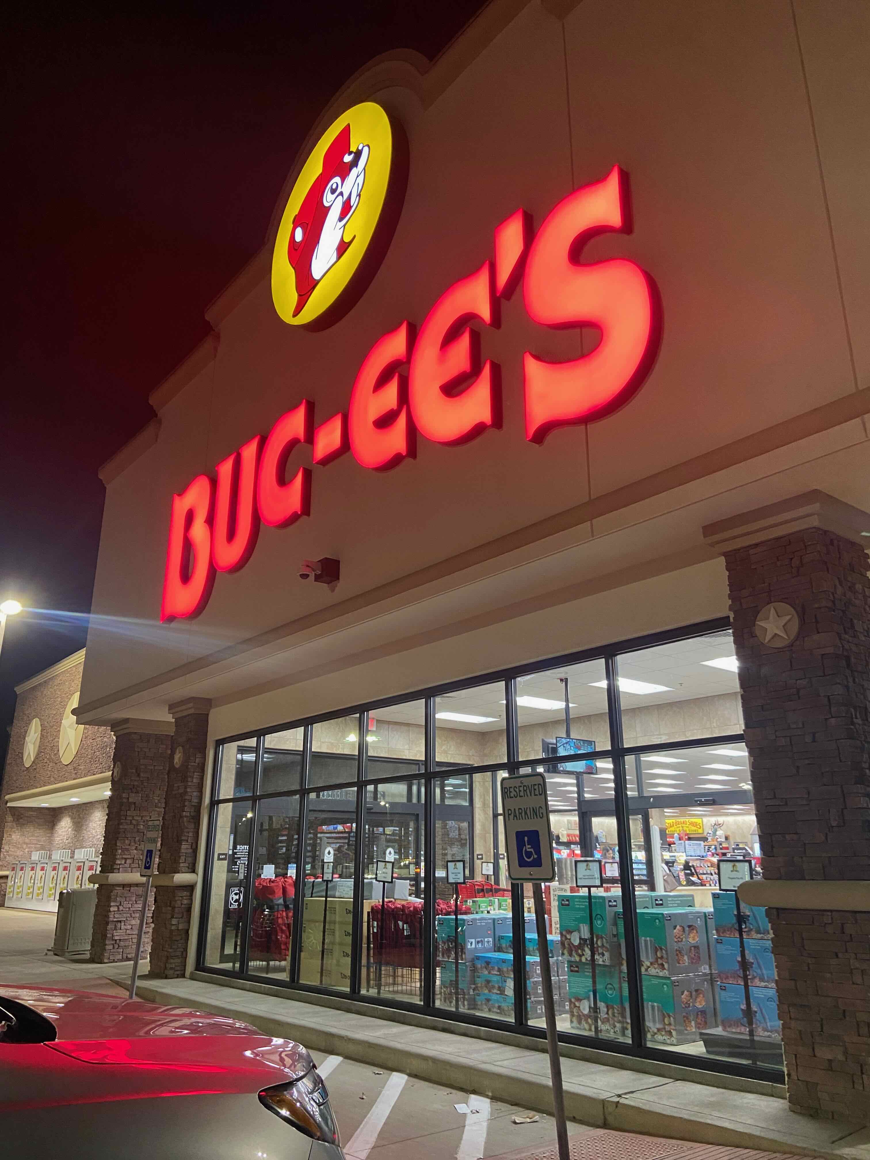 PHOTO: Entrance facade at night — the glowing red Buc-ee's sign, stone columns, warm storefront light spilling into the Texas dark