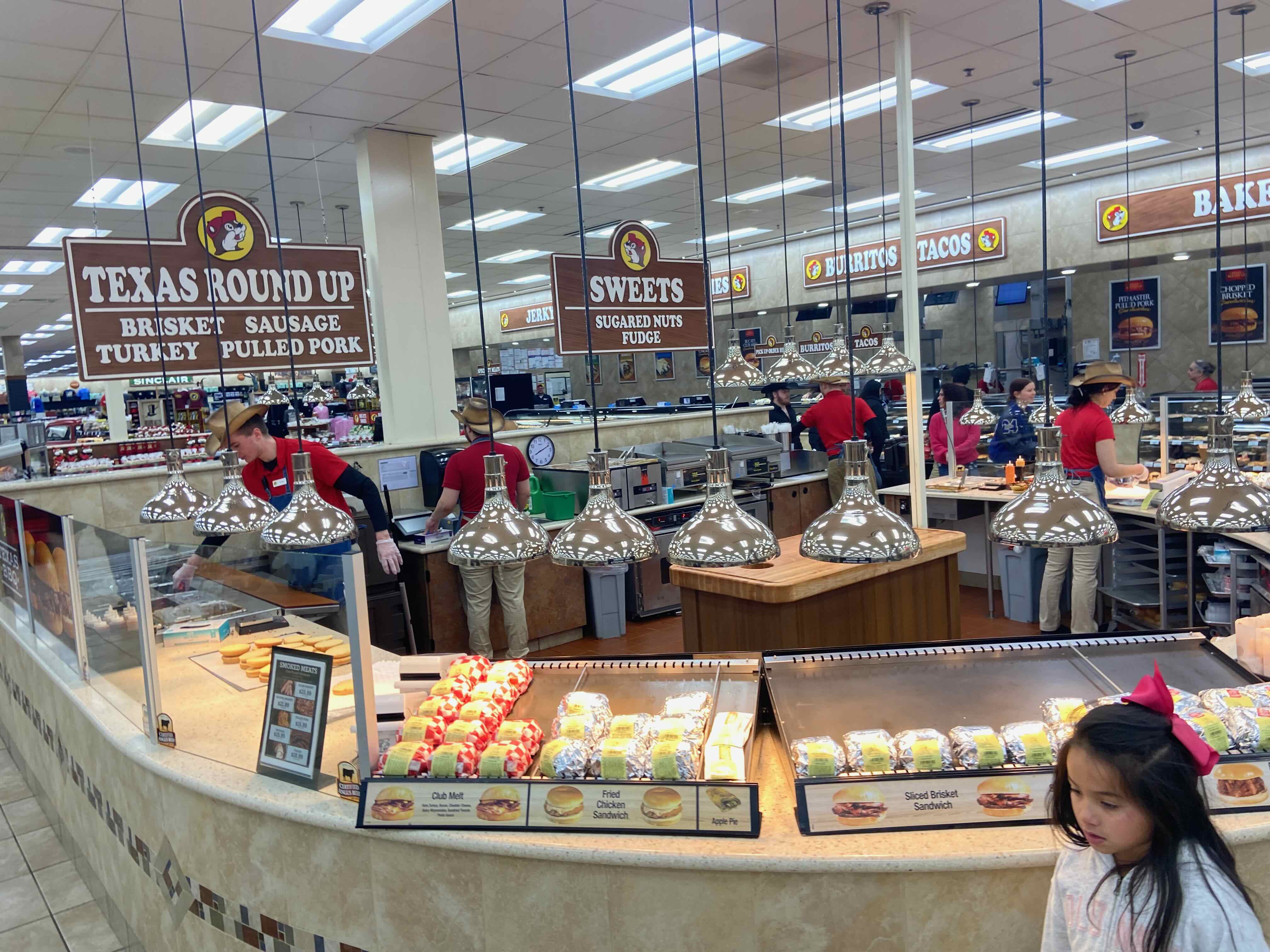 PHOTO: Food hall — "Texas Round Up," "Sweets," "Burritos & Tacos," "Bakery" signage wall with couple browsing pastry case