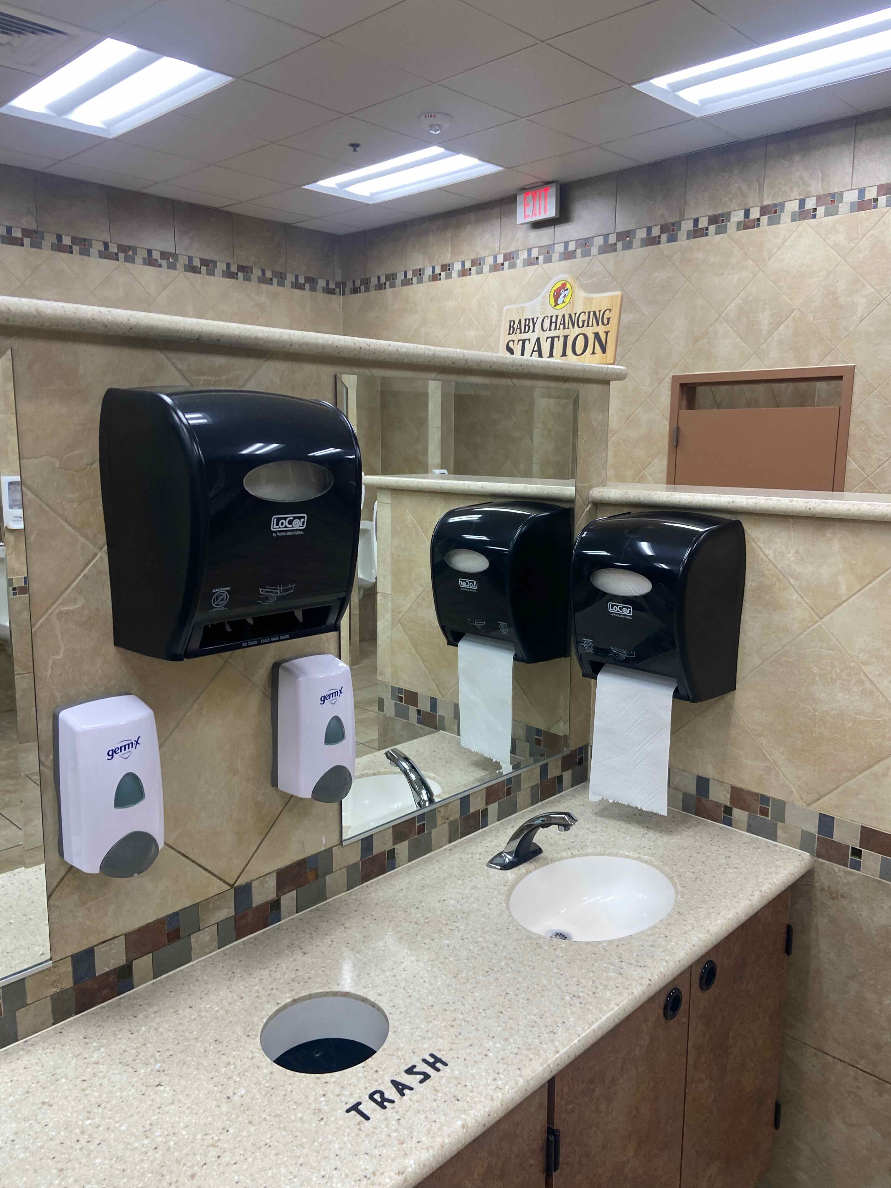 PHOTO: Sink counter — two soap dispensers, two paper towel dispensers, trash cutout labeled "TRASH" in the counter