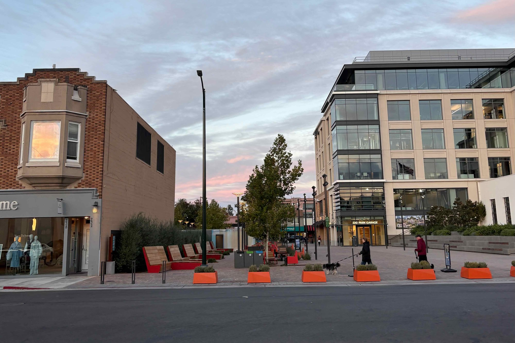 Burlingame's new Town Square at dusk: wooden lounge chairs, young trees and orange planters on a plaza built on a former downtown parking lot, with people walking a dog toward the 220 Park garage