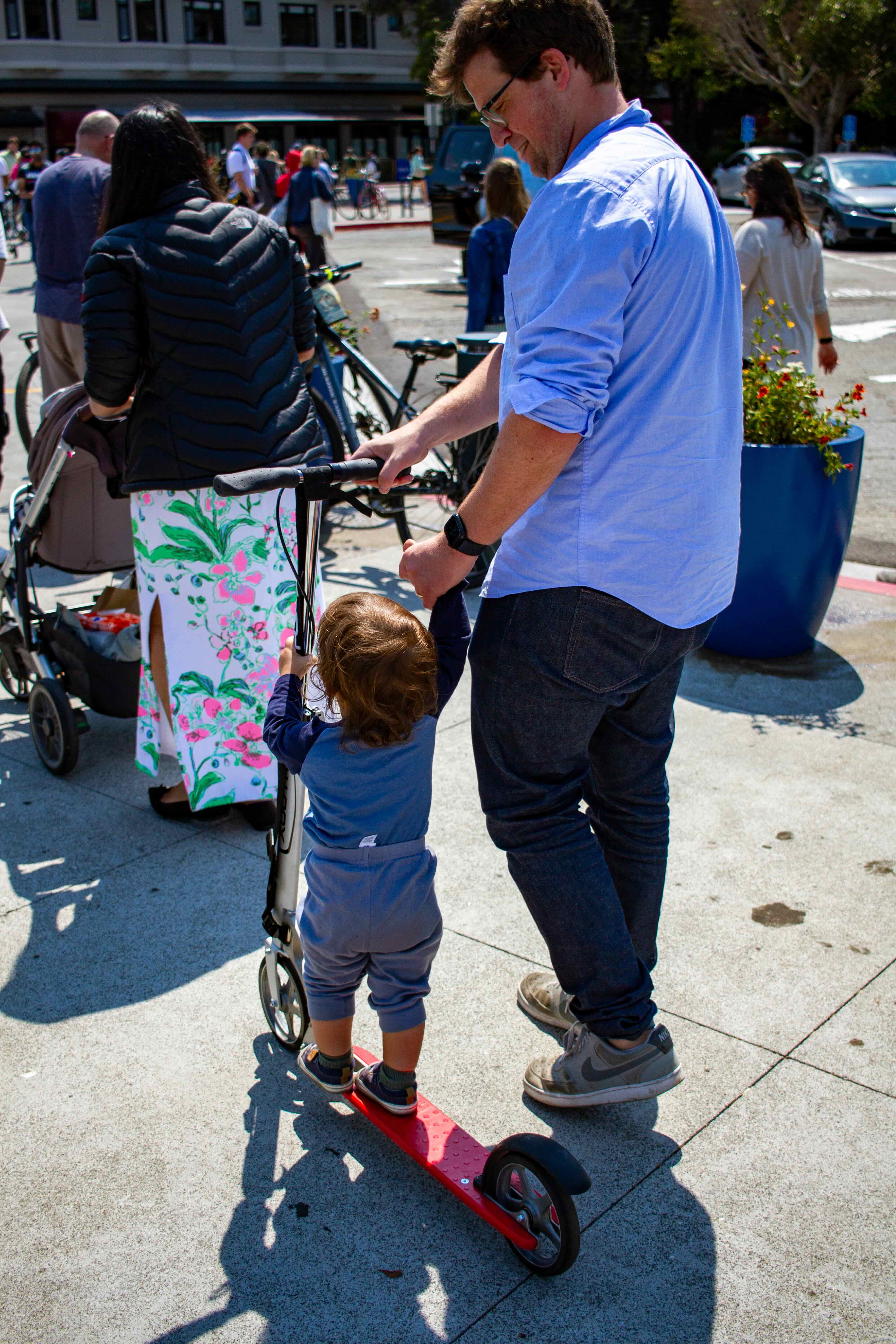 a father and son with a kickscooter, the son holding the steering column while standing on the deck and the father pushing the handlebars