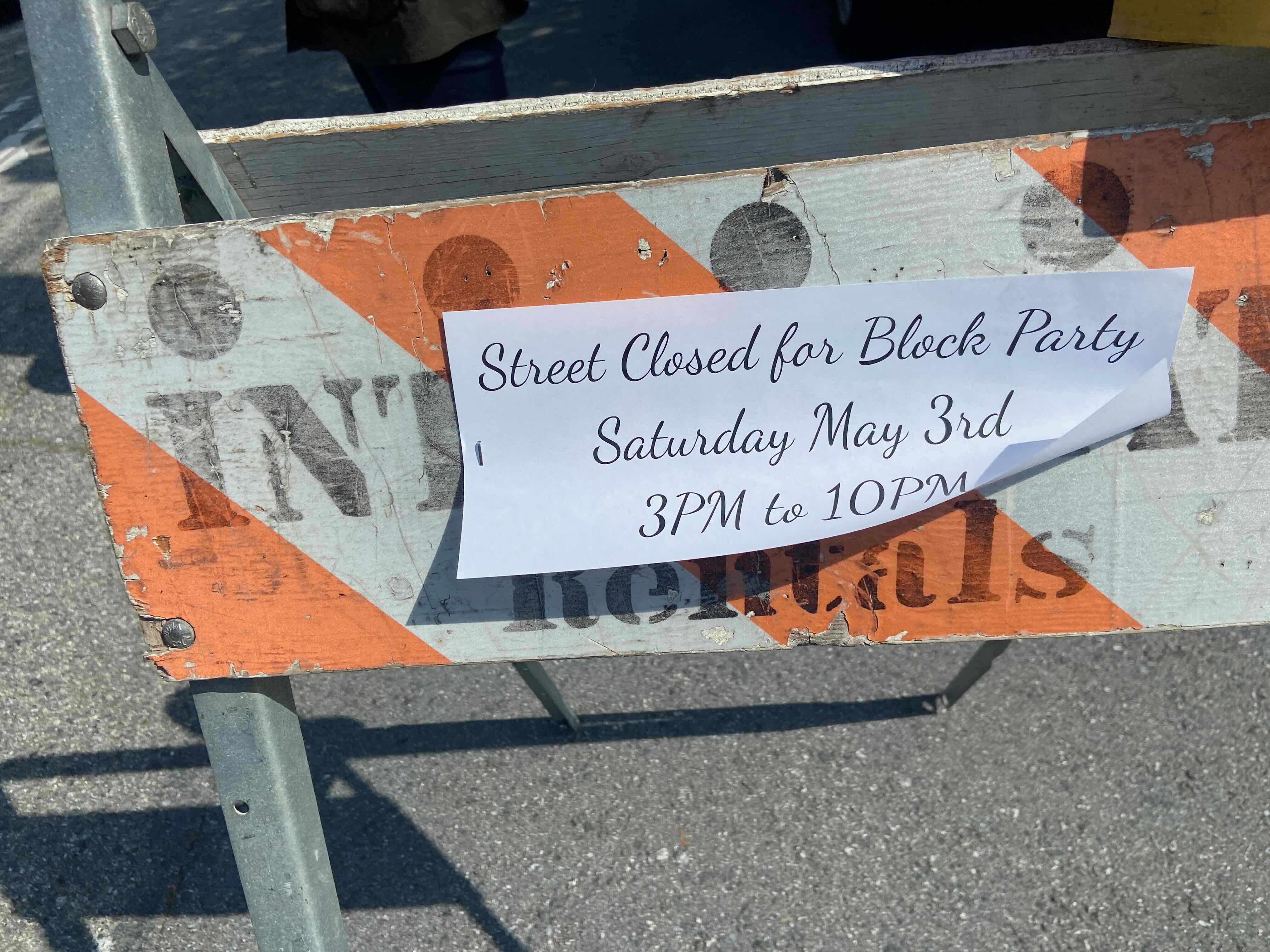 A beat-up rental barricade with a printed sign reading 'Street Closed for Block Party Saturday May 3rd 3PM to 10PM'